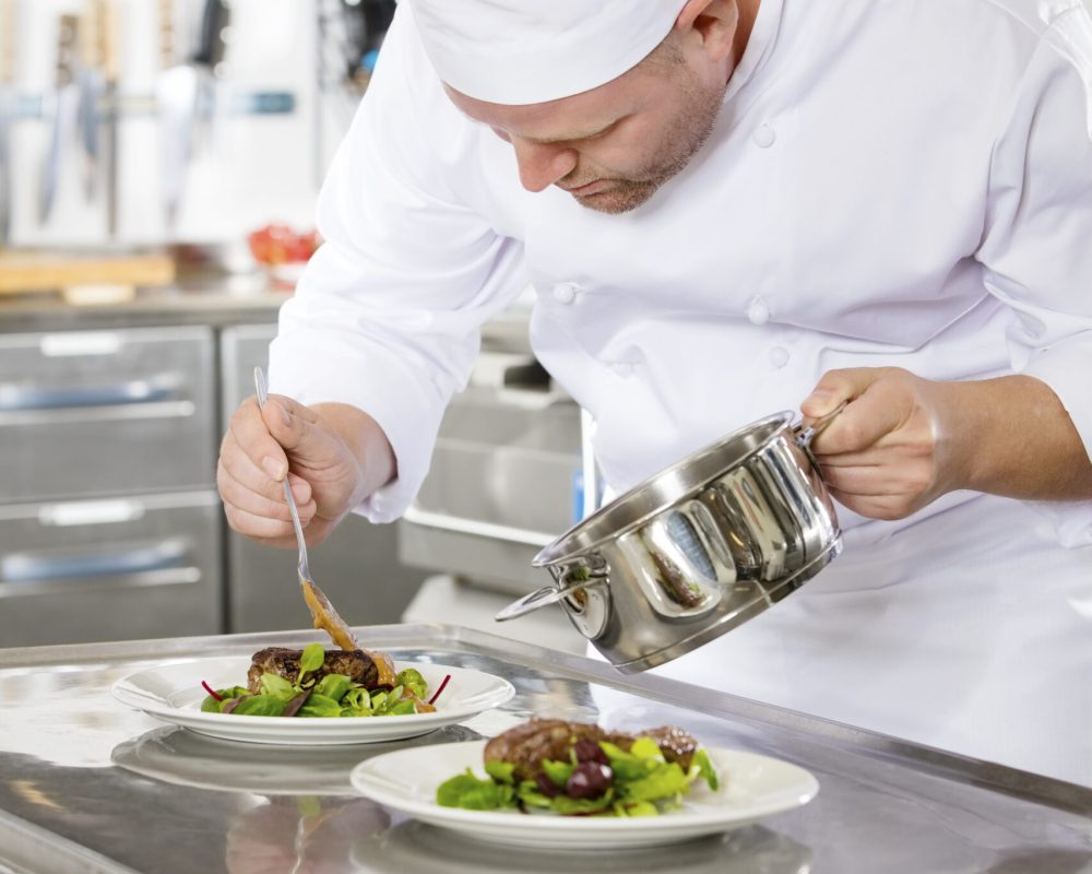 Focused chef adds gravy to a meat dish in a professional kitchen at gourmet restaurant or hotel.